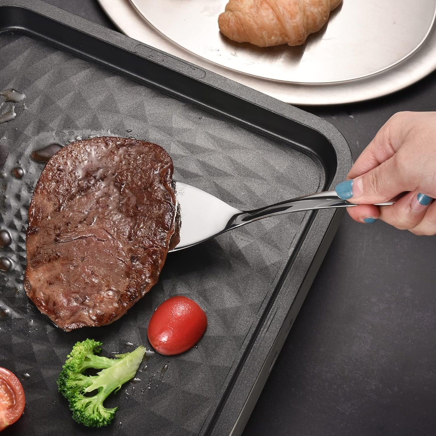 IMEEA Small Stainless Steel Turner lifting a steak from a frying pan, showcasing its practical cooking use